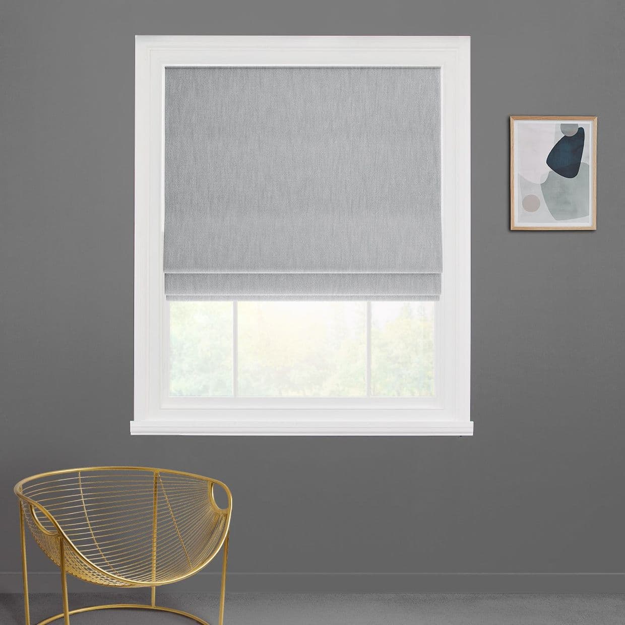 Gray fabric Roman blind fully lowered with subtle folds, filtering daylight from a white-framed window; mounted on a gray wall beside abstract framed art above a gold wire chair.