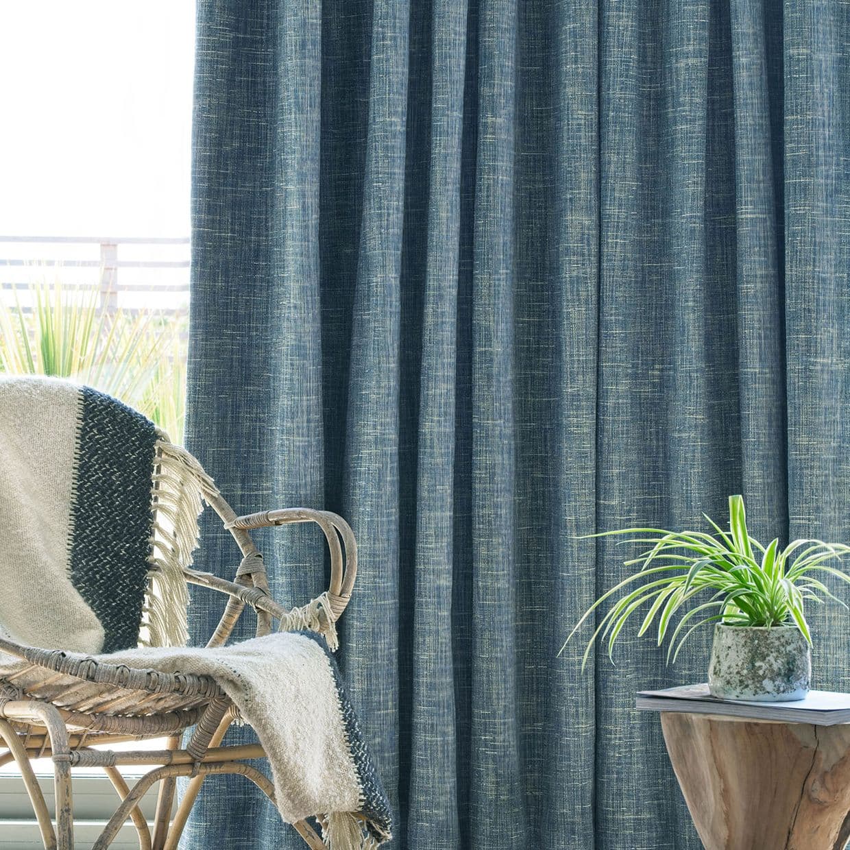 Textured teal curtains, floor-to-ceiling and mostly closed with a subtle crosshatch weave, filtering daylight; a wicker chair draped with a throw and a wooden side table holding a potted plant.