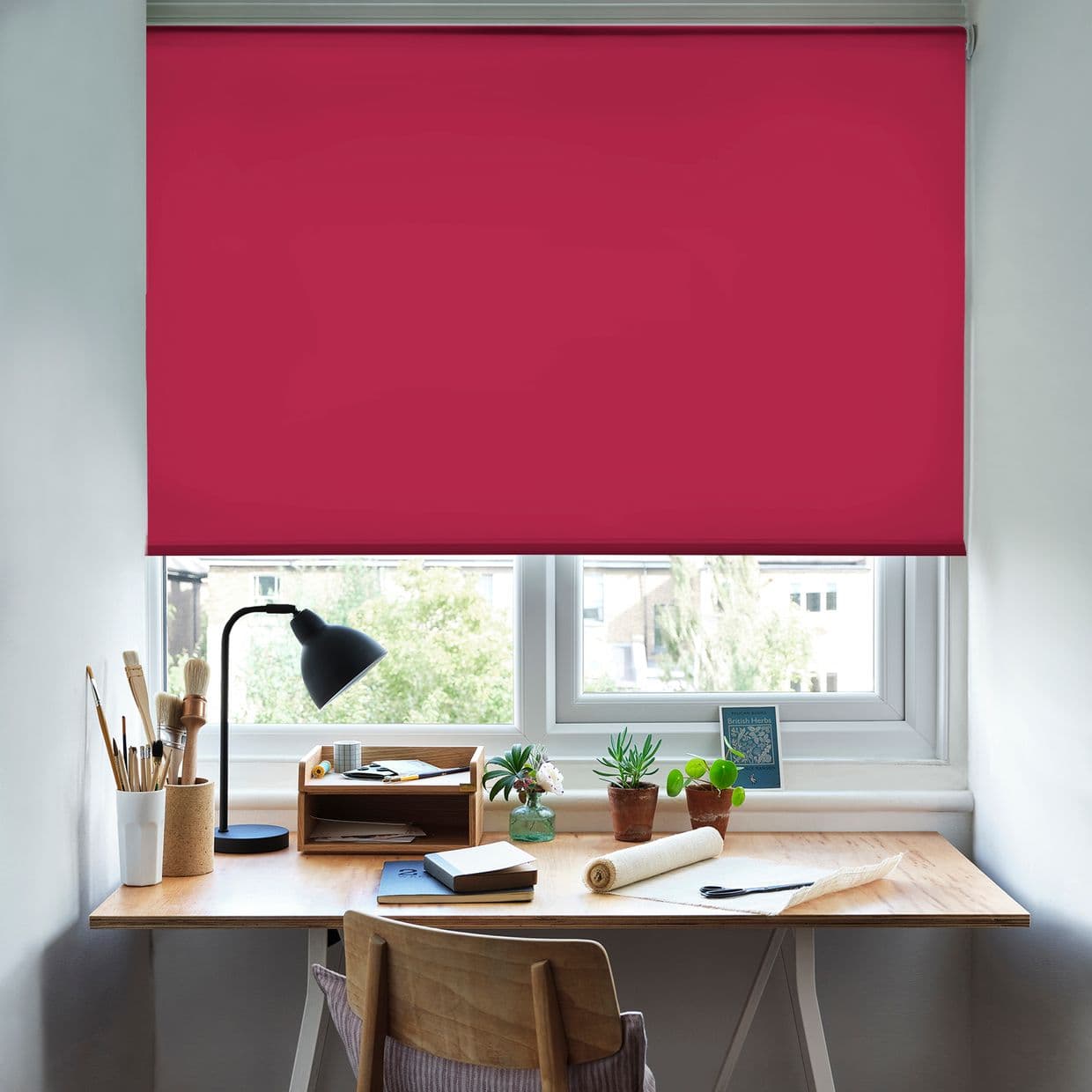 Fuchsia roller blind, fully lowered across a double window with hem; blocking most light while a narrow bottom gap admits daylight â€” above a wooden desk with lamp, plants, stationery.