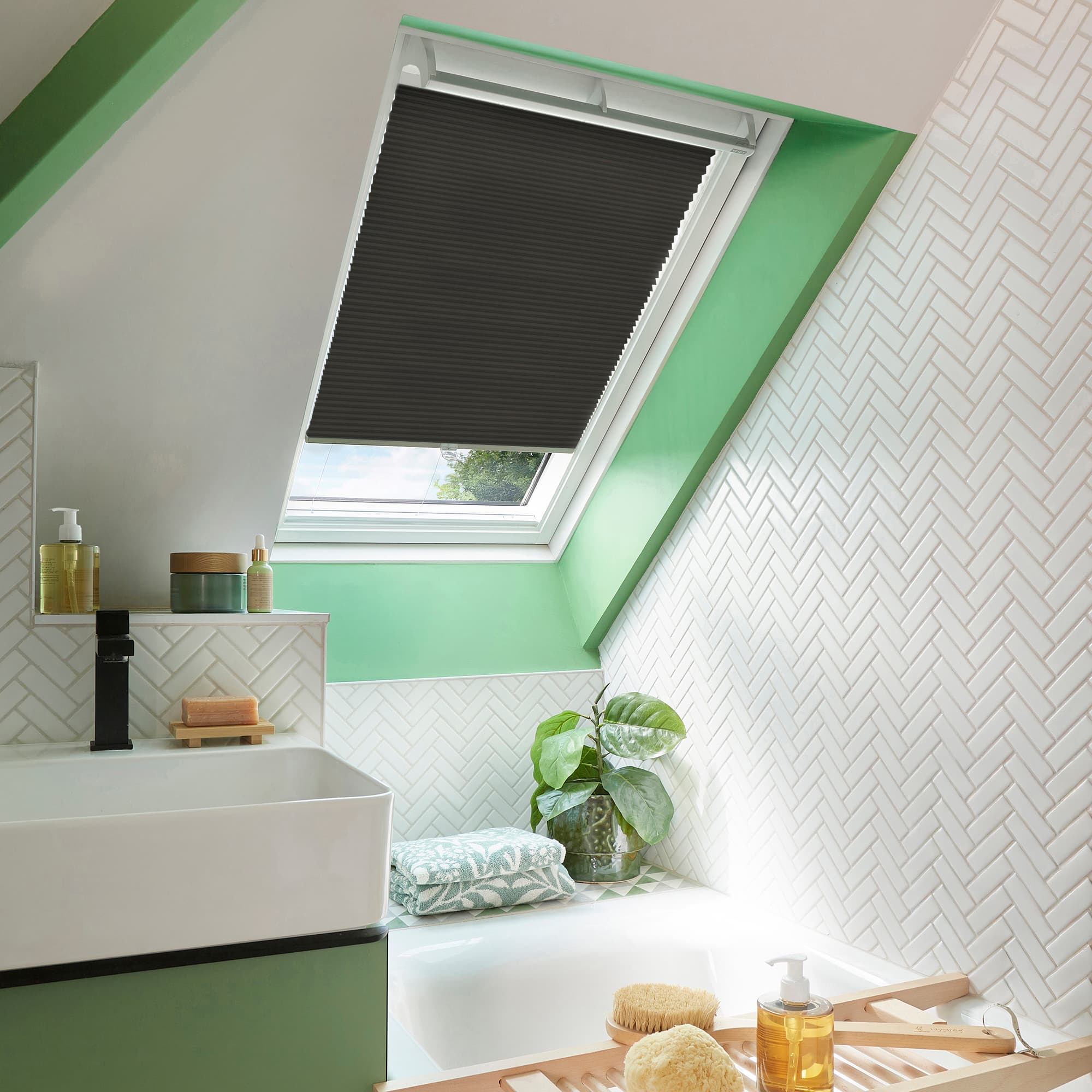 Black pleated skylight blind, partially lowered across a white roof window, blocking most light; attic bathroom with mint-green sloped walls, white herringbone tiles, sink, bathtub, plant and bath tray.