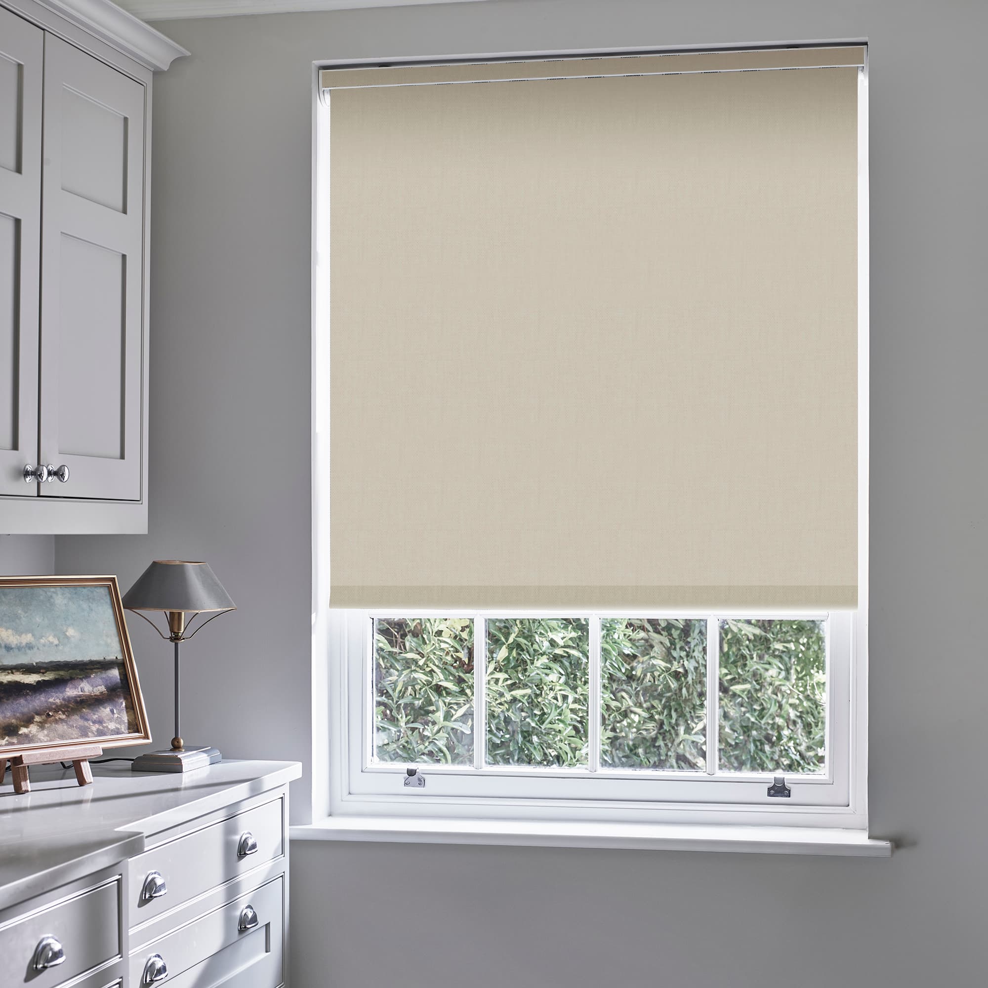 Beige roller blind lowered nearly fully, filtering light and covering the upper sash; gray-painted room with white cabinetry, lamp and painting, sash window shows leafy hedge outside.