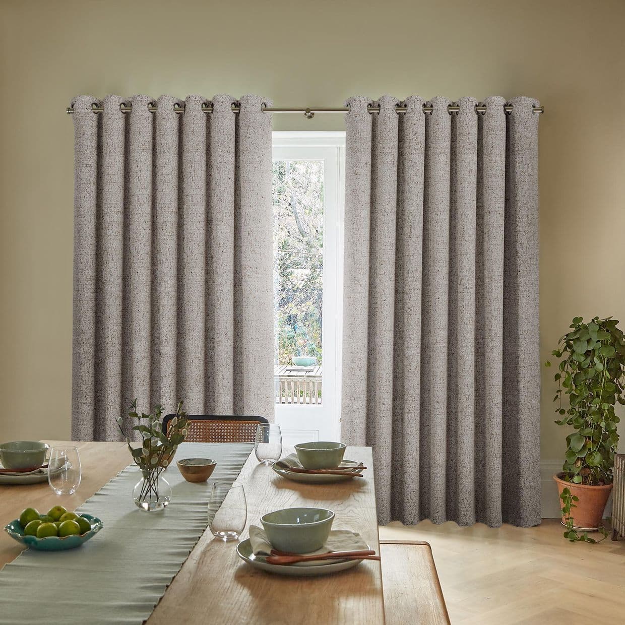 Textured gray grommet curtains on a metal rod, mostly closed with a narrow central gap filtering daylight through window; dining room with wooden table, place settings and potted plant.