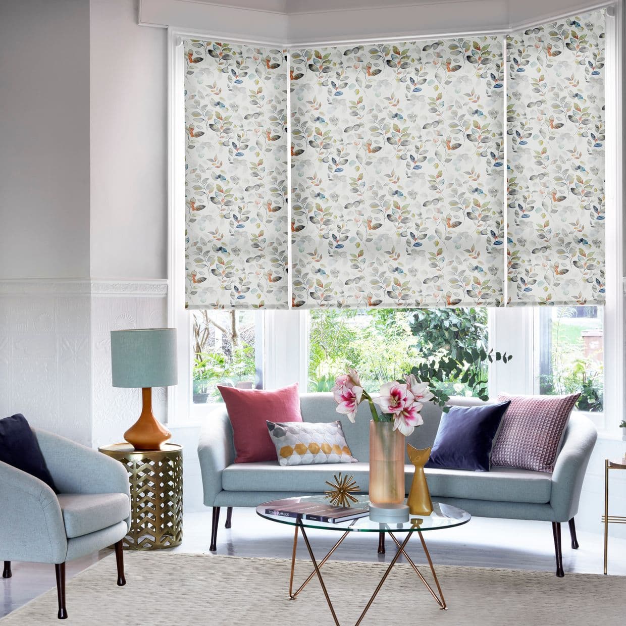 Floral leaf-patterned roller blinds, three-panel across a bay window, fully lowered, softly diffusing daylight. Living room setting with pale blue sofas, colorful cushions, glass coffee table, lamp and garden view.