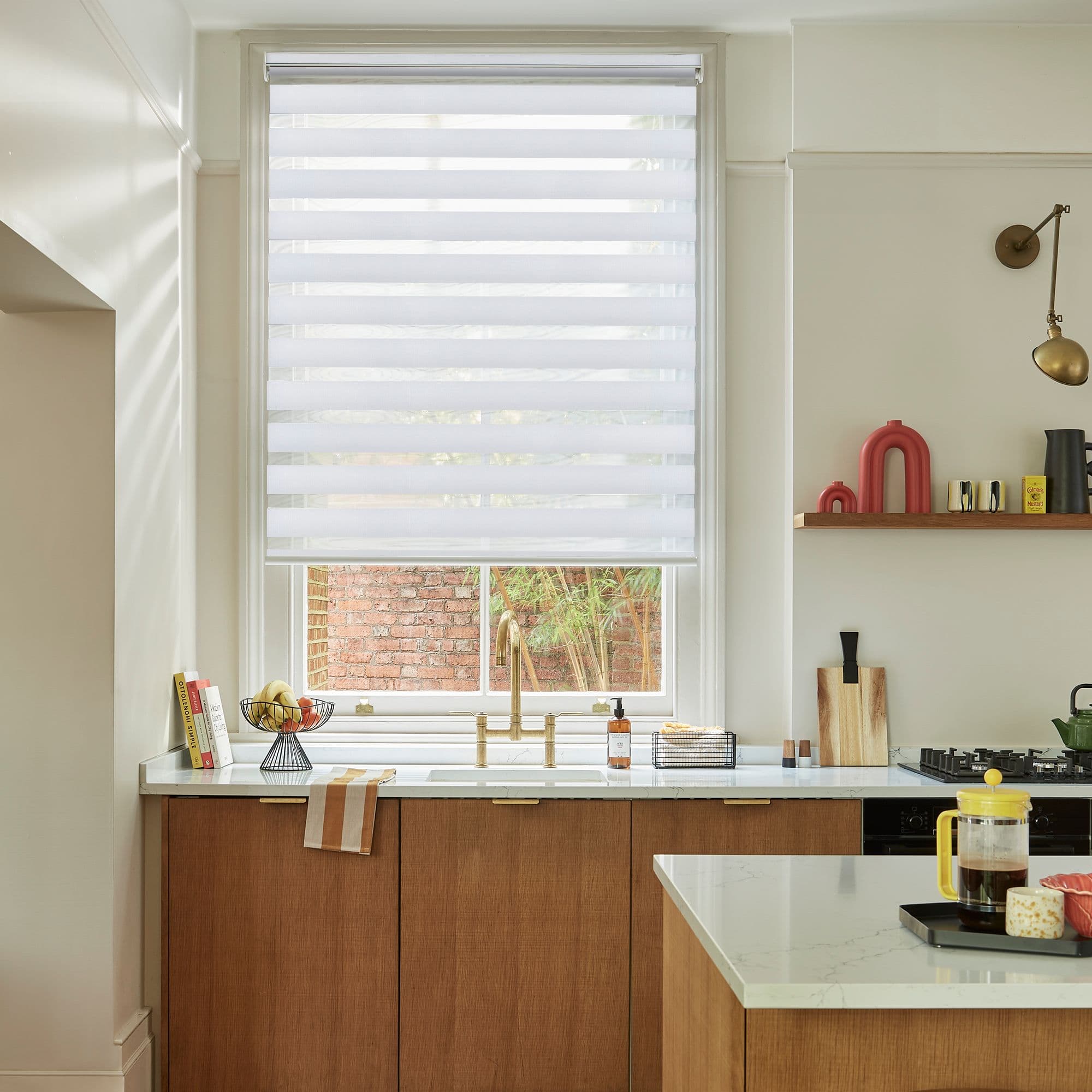 White zebra blind covering the window with alternating opaque and sheer horizontal stripes, filtering daylight; above a kitchen sink with brass faucet, marble counters, wooden cabinets, and a brick exterior.