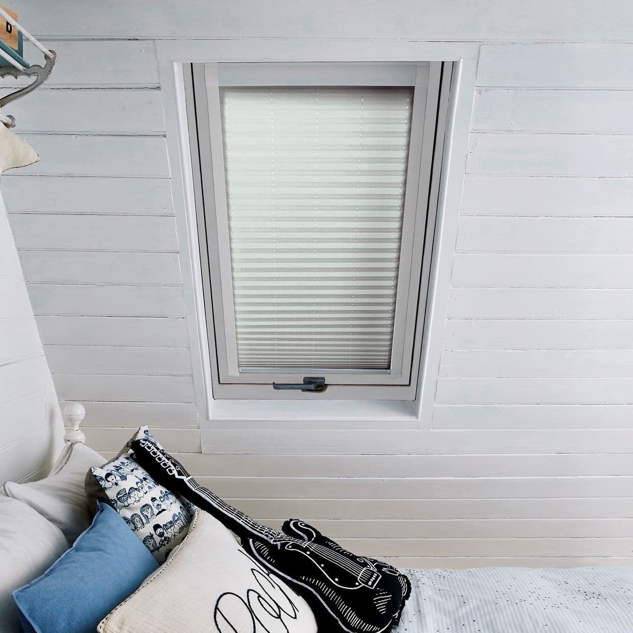 Pleated white blind in a recessed skylight, fully lowered and closed, diffusing soft daylight into a white-painted attic bedroom above a bed with blue patterned pillows and a guitar-shaped cushion.