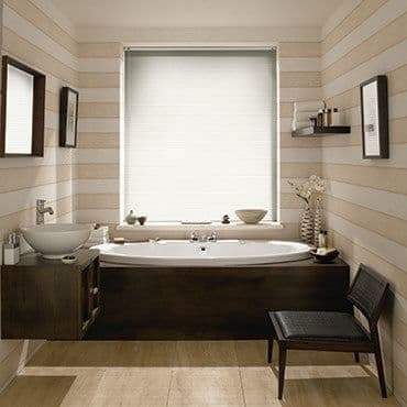 White pleated roller blind fully lowered over window, diffusing soft daylight; below, an alcove bathtub set in dark wood surround, beige horizontal-striped walls, floating shelf with toiletries, woven chair.