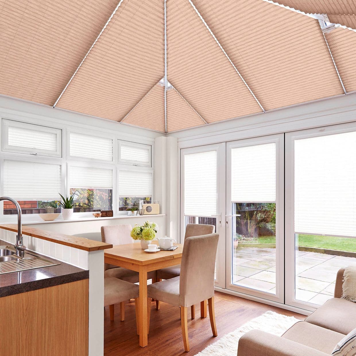Beige pleated roof blinds cover the pitched conservatory ceiling, with white pleated blinds on windows and French doors; filtering soft daylight into a bright kitchen-dining space overlooking a patio garden.