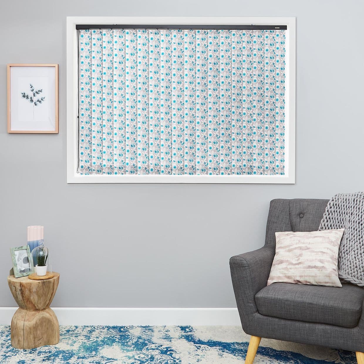 Patterned fabric vertical blinds (blue floral), fully closed and filtering light inside a white-framed window, in a grey living room with armchair, side table, print and blue rug.
