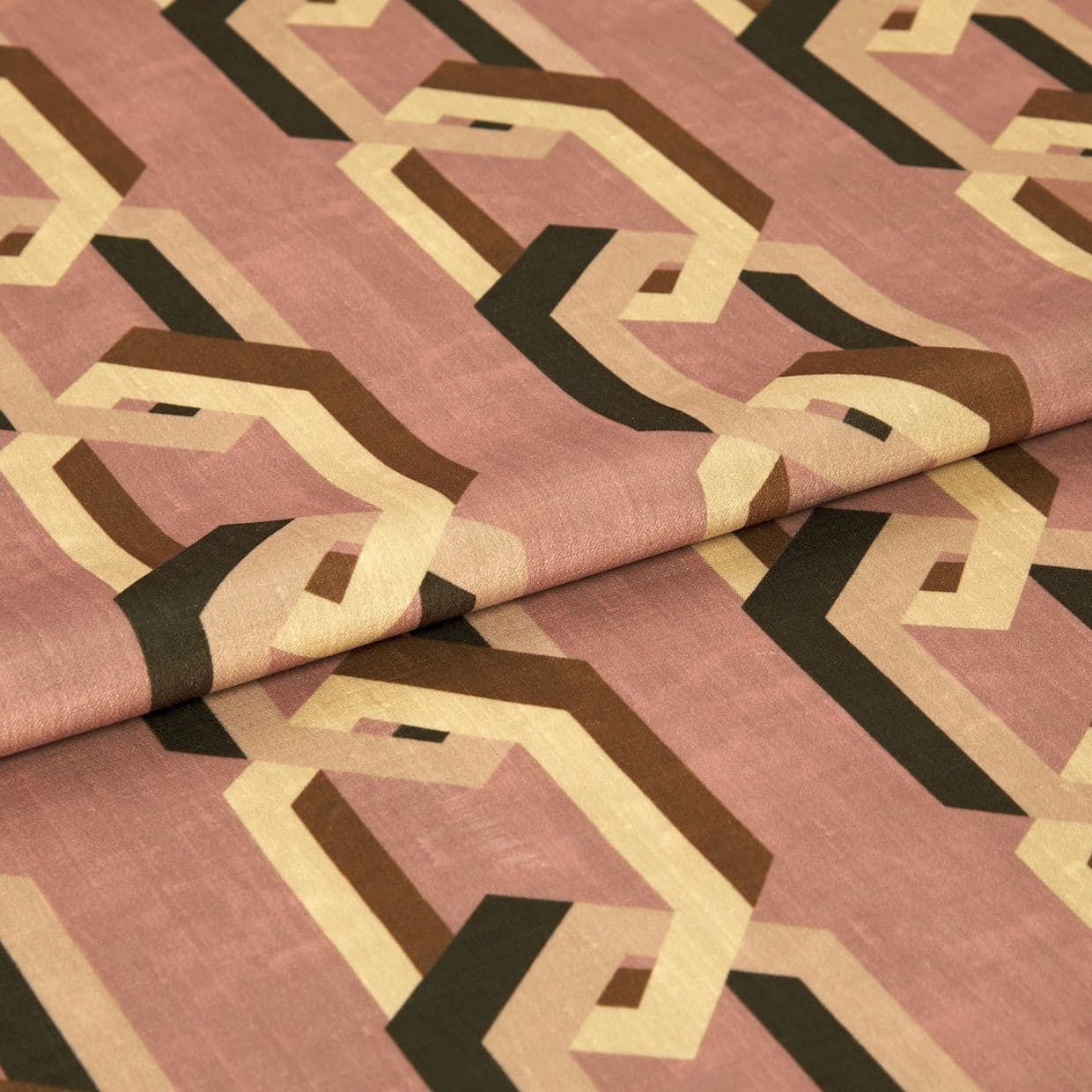 Geometric-patterned fabric with interlocking beige, brown and black hexagonal chains on a dusty rose background, folded across the center, lying on a flat surface under soft, neutral indoor lighting.