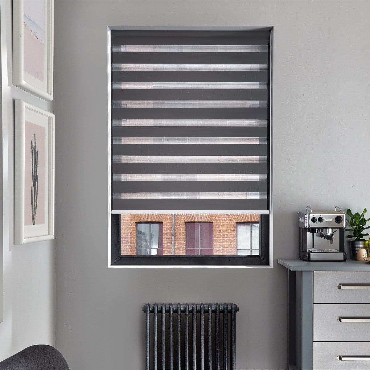 Grey dual-layer roller blind with alternating opaque and sheer horizontal stripes, lowered to filter light while partially revealing the lower sash; small kitchen corner, espresso machine, radiator, brick building outside.
