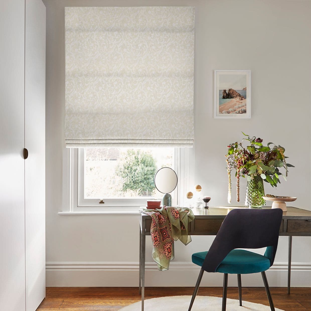 Damask cream Roman blind, fully lowered with horizontal folds, diffusing daylight; over a sash window above a desk in a neutral-lit home office with teal chair, mirror and vase of flowers.