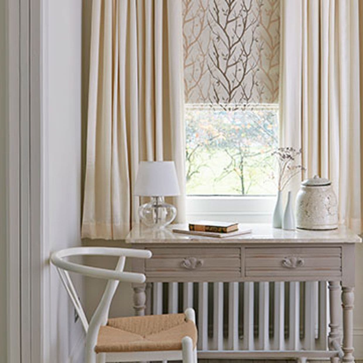Cream pleated curtains framing a patterned Roman shade with branch motif; shade partially lowered, filtering soft daylight onto a pale wooden desk with lamp, chair, vases beside a garden view.