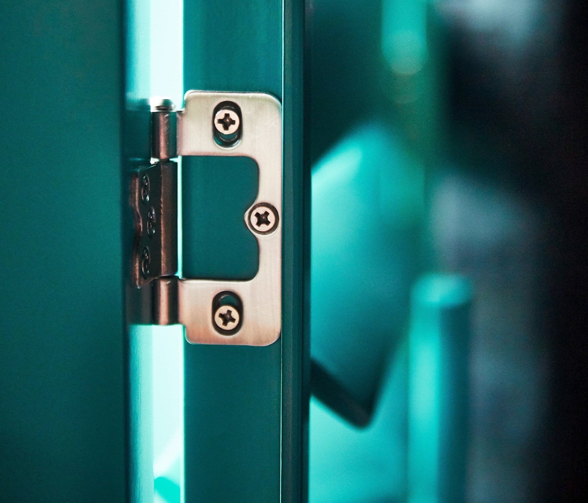 A stainless-steel door hinge mounted on a teal door edge, holding the door slightly ajar; indoor scene with cool turquoise lighting, shallow depth of field and a softly blurred background.