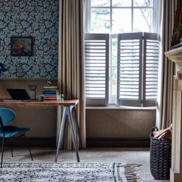 Beige floor-length curtains frame a tall window; white plantation shutters cover the lower sash with tilted horizontal slats, partially open and filtering daylight into a cozy studyâ€”desk and patterned wallpaper.
