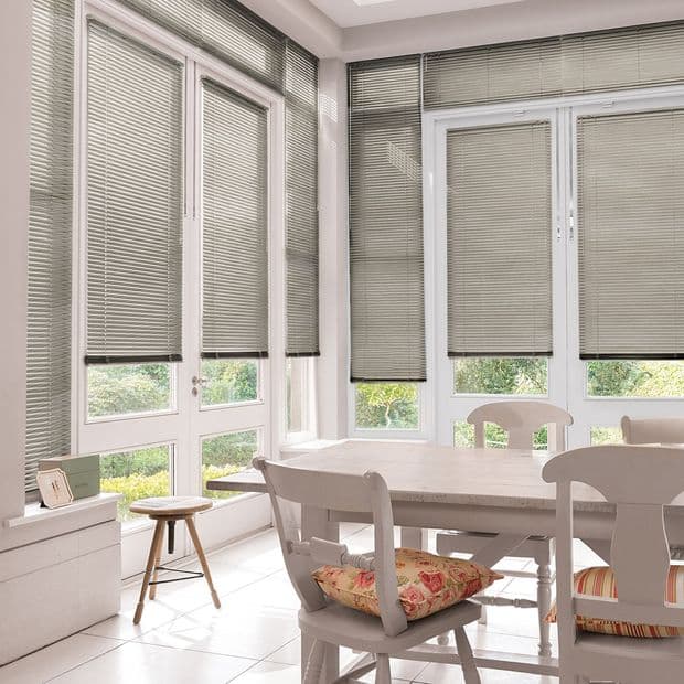 Gray horizontal Venetian blinds covering multiple tall windows and French doors, lowered and tilted to filter daylight; bright dining/sunroom with white table, chairs, cushions and garden view beyond.