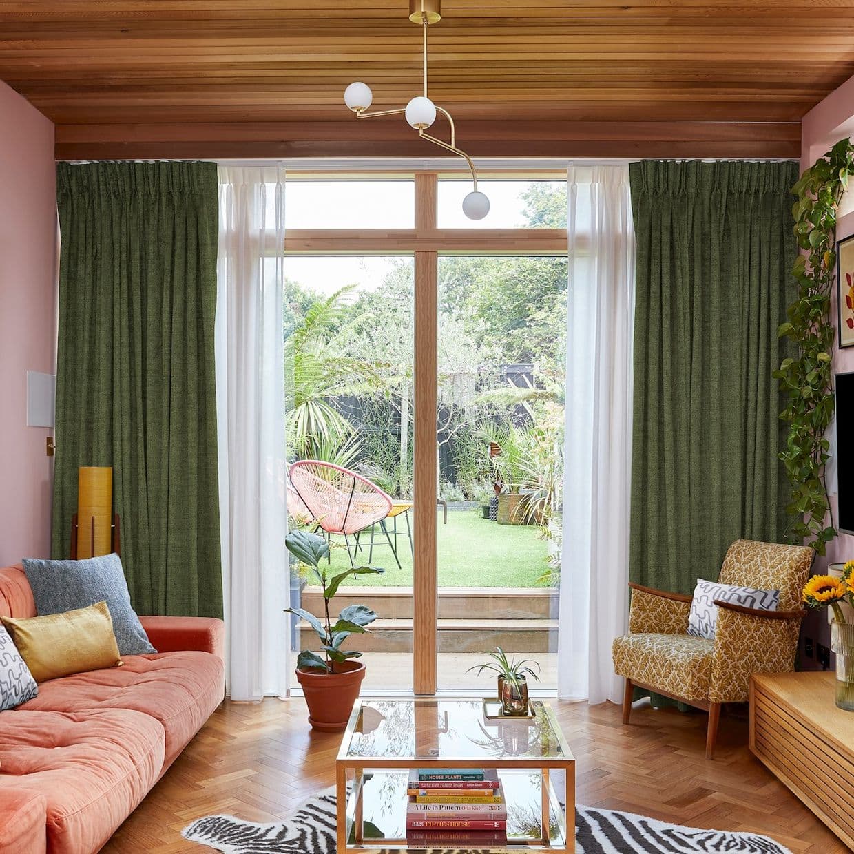 Green pleated curtains with white sheers, hung either side of a wooden-framed glass door, framing and filtering daylight; midcentury living room with coral sofa, patterned armchair and garden view.