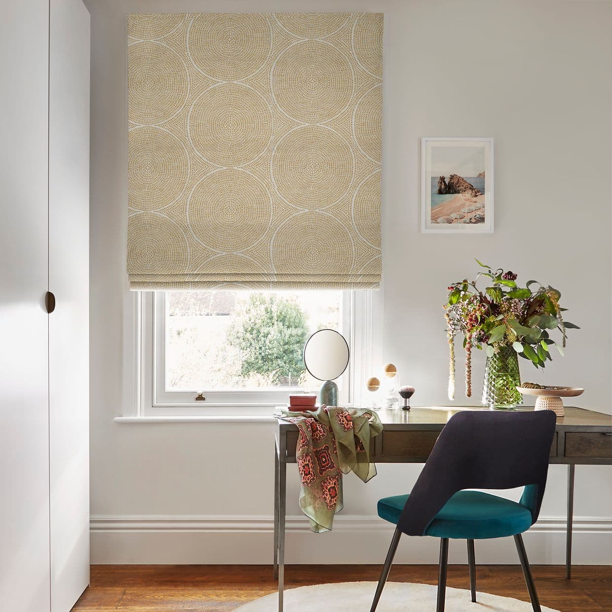 Beige patterned Roman blind, lowered over the upper sash with concentric circular motifs, filtering soft daylight; above a small desk/vanity with teal chair, round mirror, vase of flowers and garden view.