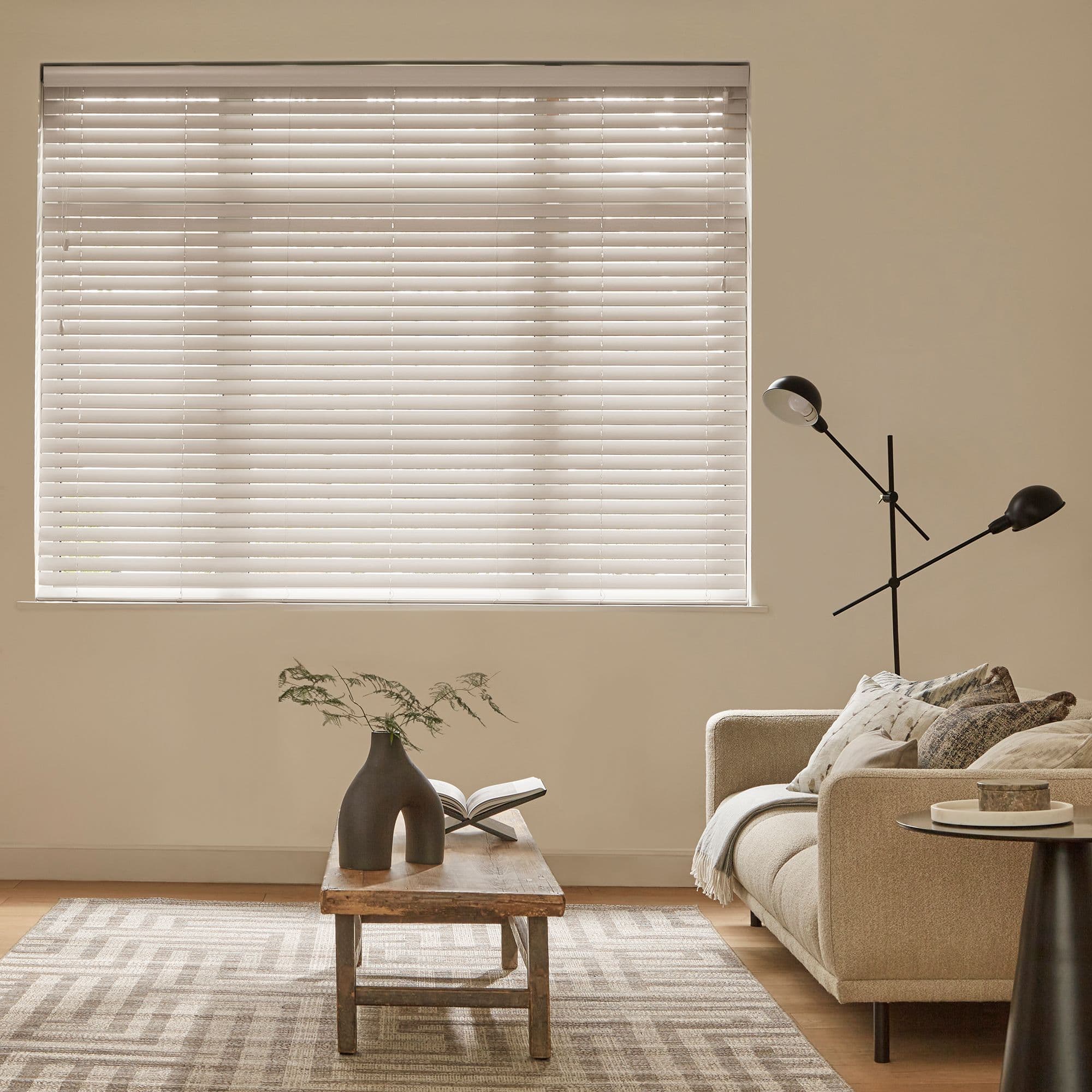 White horizontal blinds covering a large window, fully closed and softly diffusing daylight; neutral living room with beige sofa, wooden coffee table, sculptural vase, black floor lamp, patterned rug.