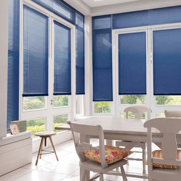 Blue horizontal Venetian blinds covering tall white-framed windows and doors, mostly lowered and filtering daylight; bright sunlit dining nook with a white table, chairs, floral cushion, stool and garden view.