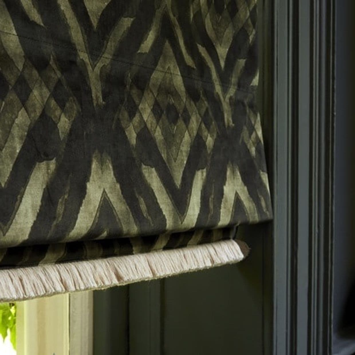 A patterned fabric Roman shade with olive-and-black geometric/ikat design and pale fringe trim hangs partially lowered and folded, covering a window within dark green trim, softly lit from the left.