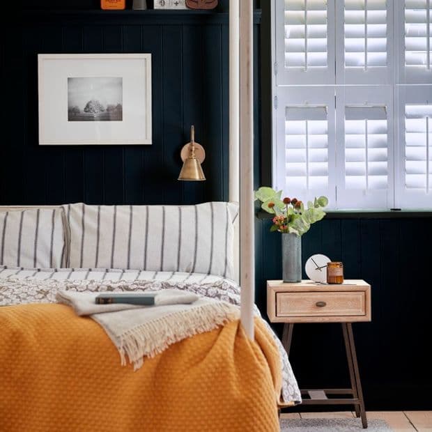 White plantation shutters covering a window, closed with louvers tilted to filter soft daylight; against dark paneled walls beside a four-post bed with striped bedding, mustard throw and wooden nightstand.