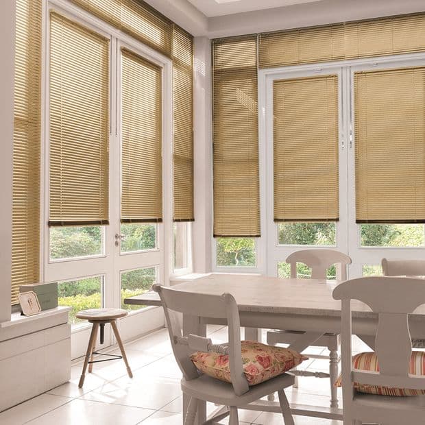 Beige horizontal Venetian blinds covering floor-to-ceiling French doors and tall windows, lowered and filtering soft daylight; dining table and chairs in a bright sunroom overlooking a green garden.