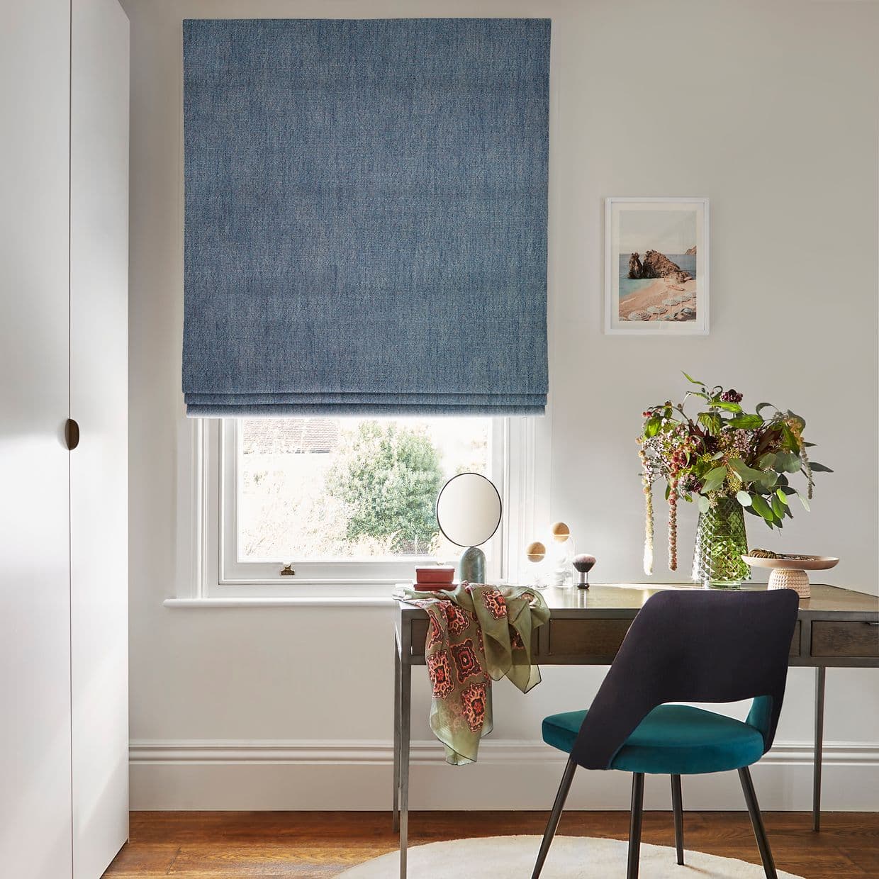 Blue textured Roman blind, lowered, blocking most daylight, leaving a light strip at the sill; over a sash window above a desk in a study with a teal chair.