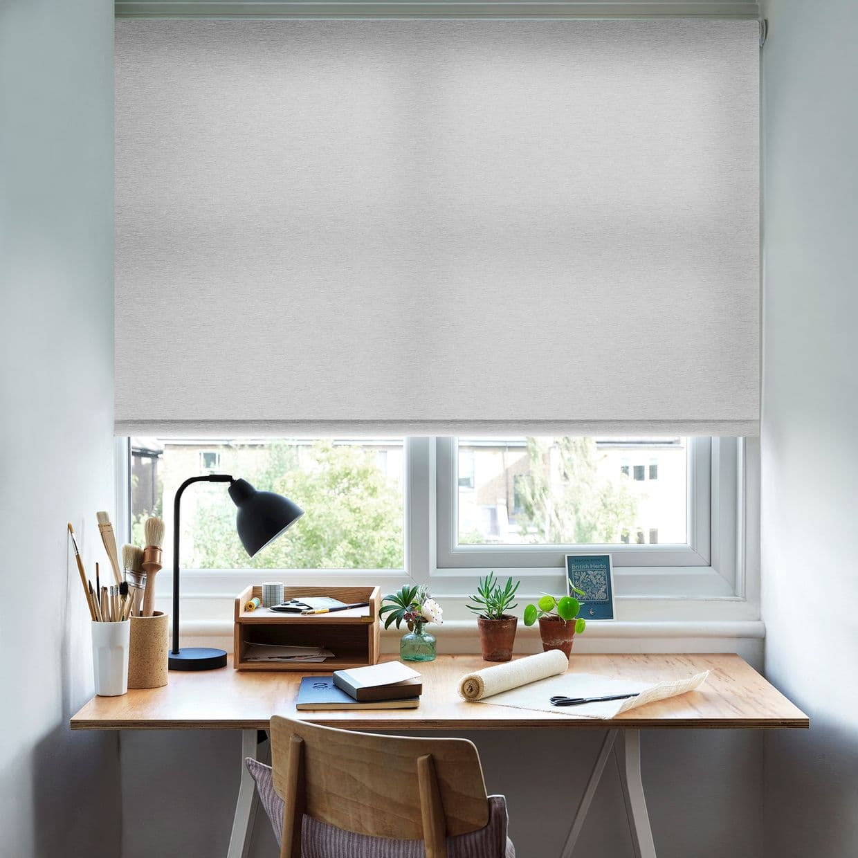 Light-gray roller blind lowered across the window, textured and slightly translucent; filtering daylight with a narrow gap at the bottom; small home workspace with wooden desk, lamp, art supplies, potted plants.