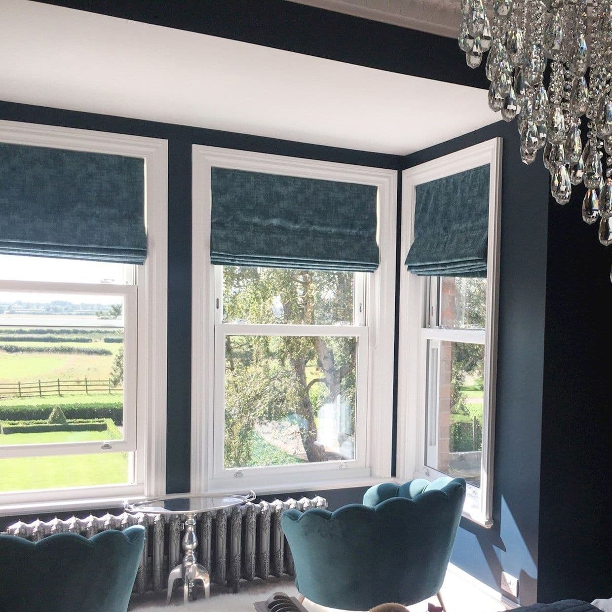 Three teal Roman blinds, half-lowered across a bay of white-framed windows, filtering sunlight into a dark-blue sitting area with teal armchairs, radiator and chandelier, view of green fields.