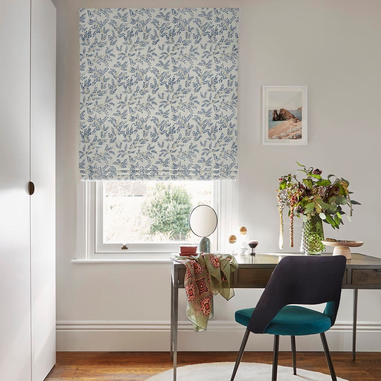 Blue-and-white floral Roman blind lowered over window, partially filtering daylight; desk with teal chair, small mirror and vase of flowers in a softly lit neutral study with framed seaside print.