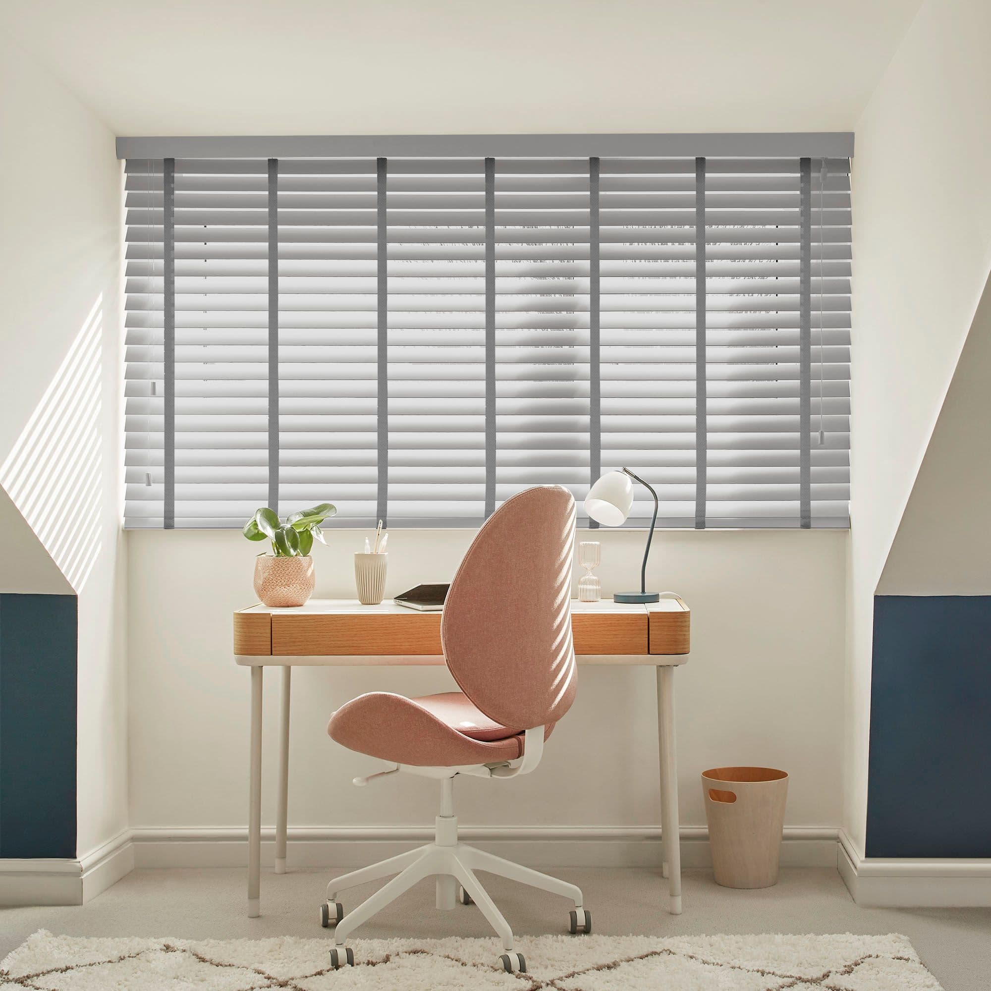 Grey horizontal Venetian blinds covering a wide skylight window, closed with slats angled to filter sunlight into an attic office; wooden desk, pink swivel chair, lamp, plant, soft daylight.