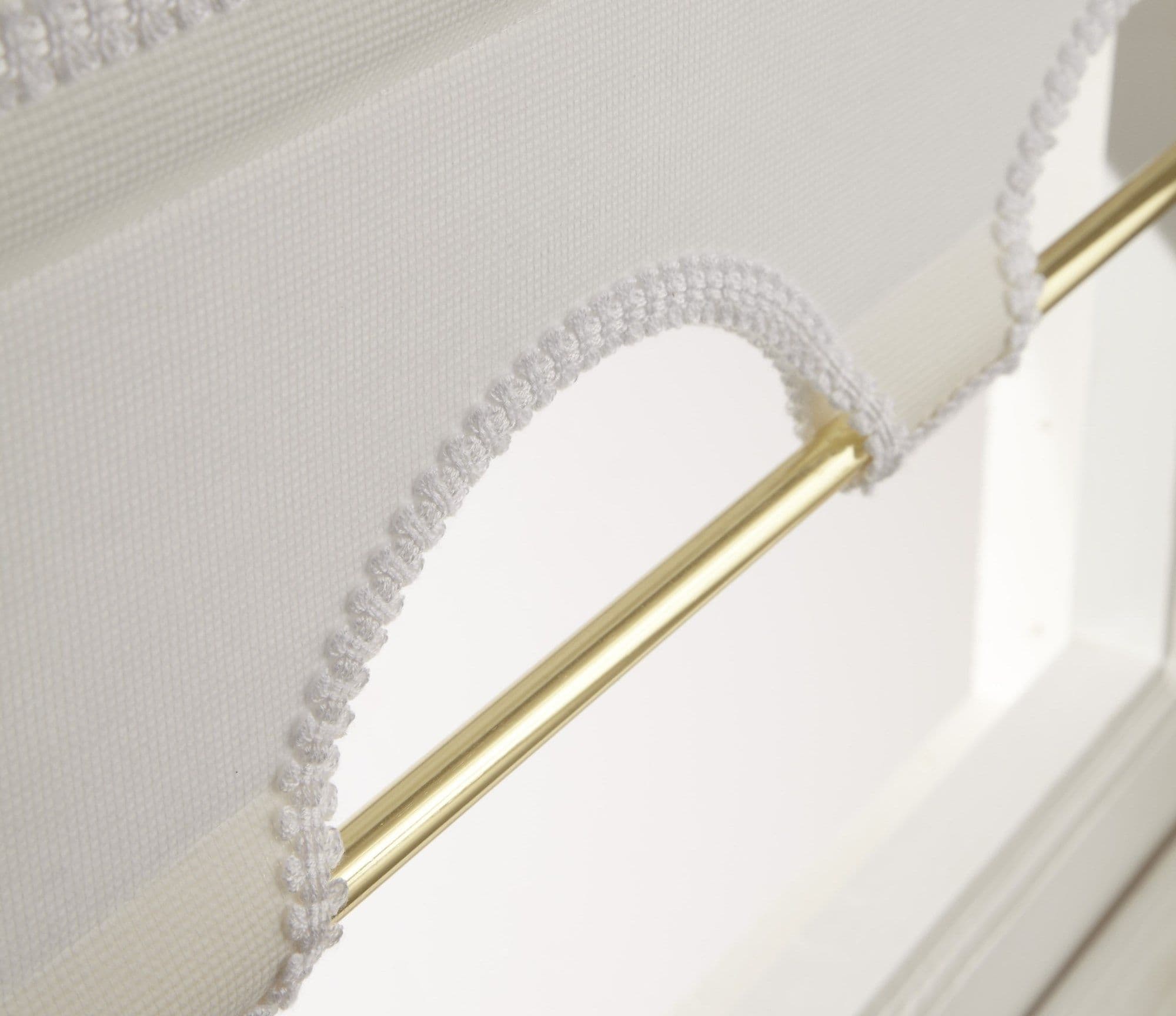 White scallopedâ€‘trim fabric blind looped over a polished brass rod, crocheted edging forming decorative arches; partially raised, filtering soft daylight above white window frame in a bright room.