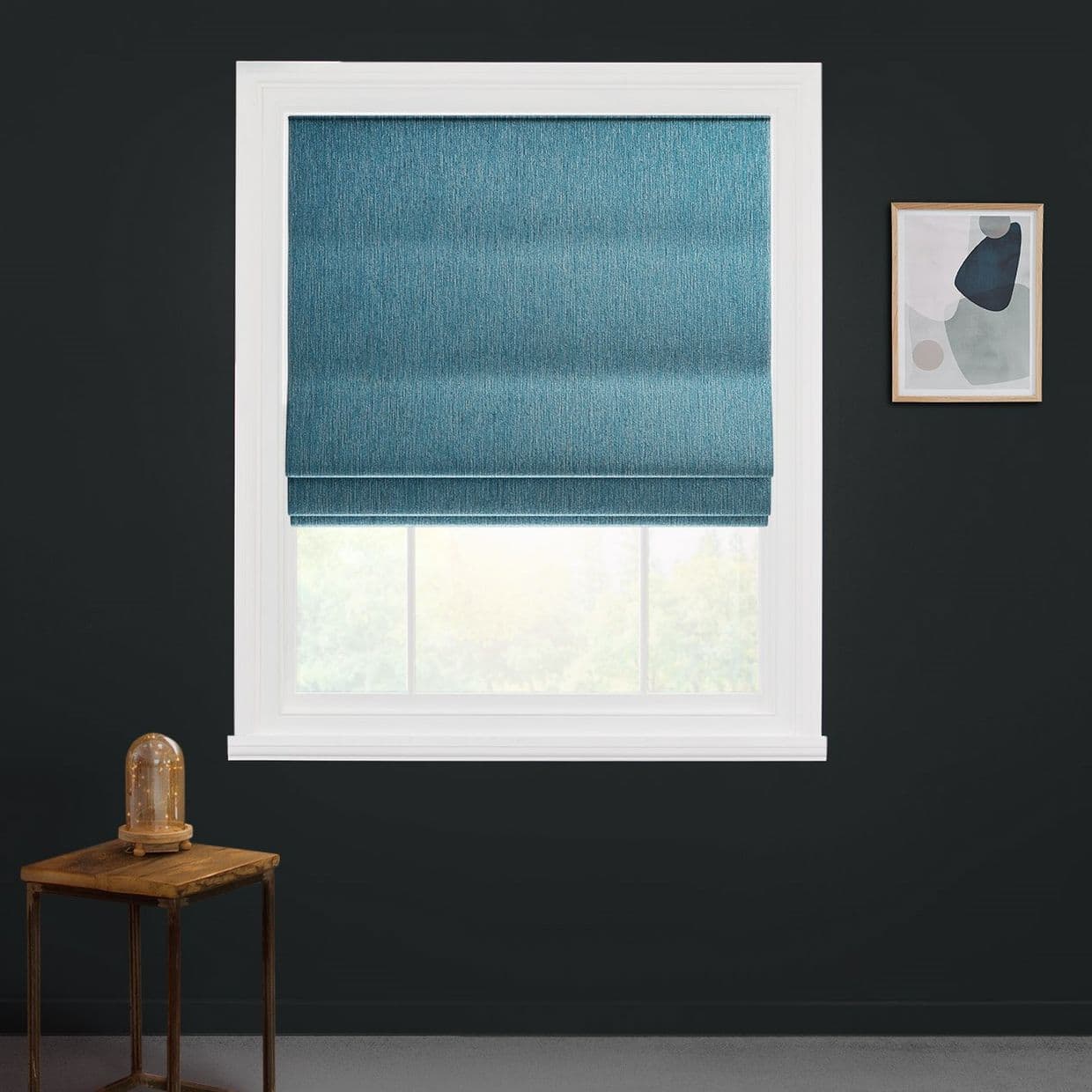 Teal fabric Roman blind, lowered mid-window with visible horizontal folds, filtering daylight; set in a white-framed window on a dark wall, beside small wooden side table and framed abstract art.