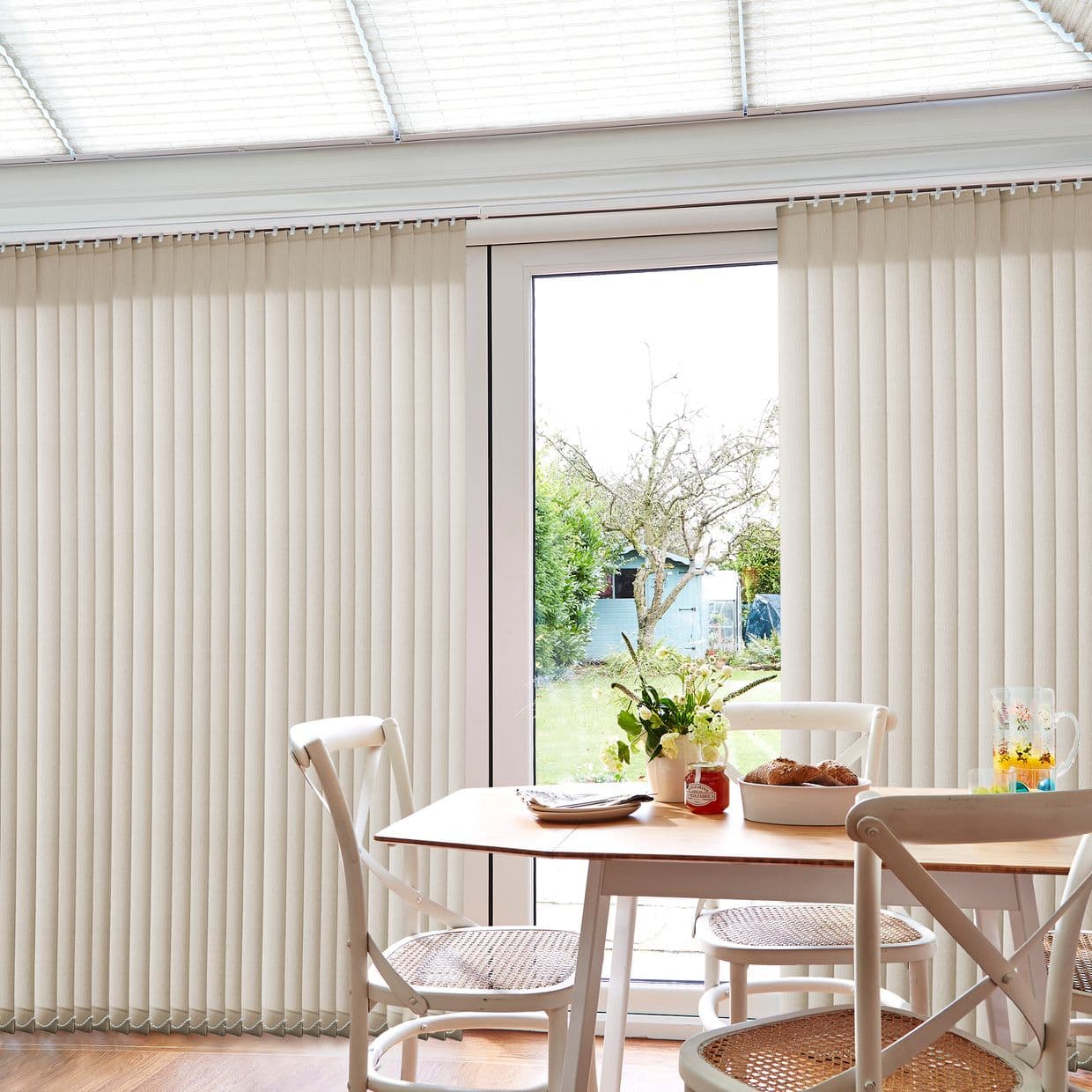 Beige vertical blinds, ceiling-mounted with pleated slats, partially parted to reveal a glass patio door, filtering soft daylight across a bright dining area with wooden table and chairs.