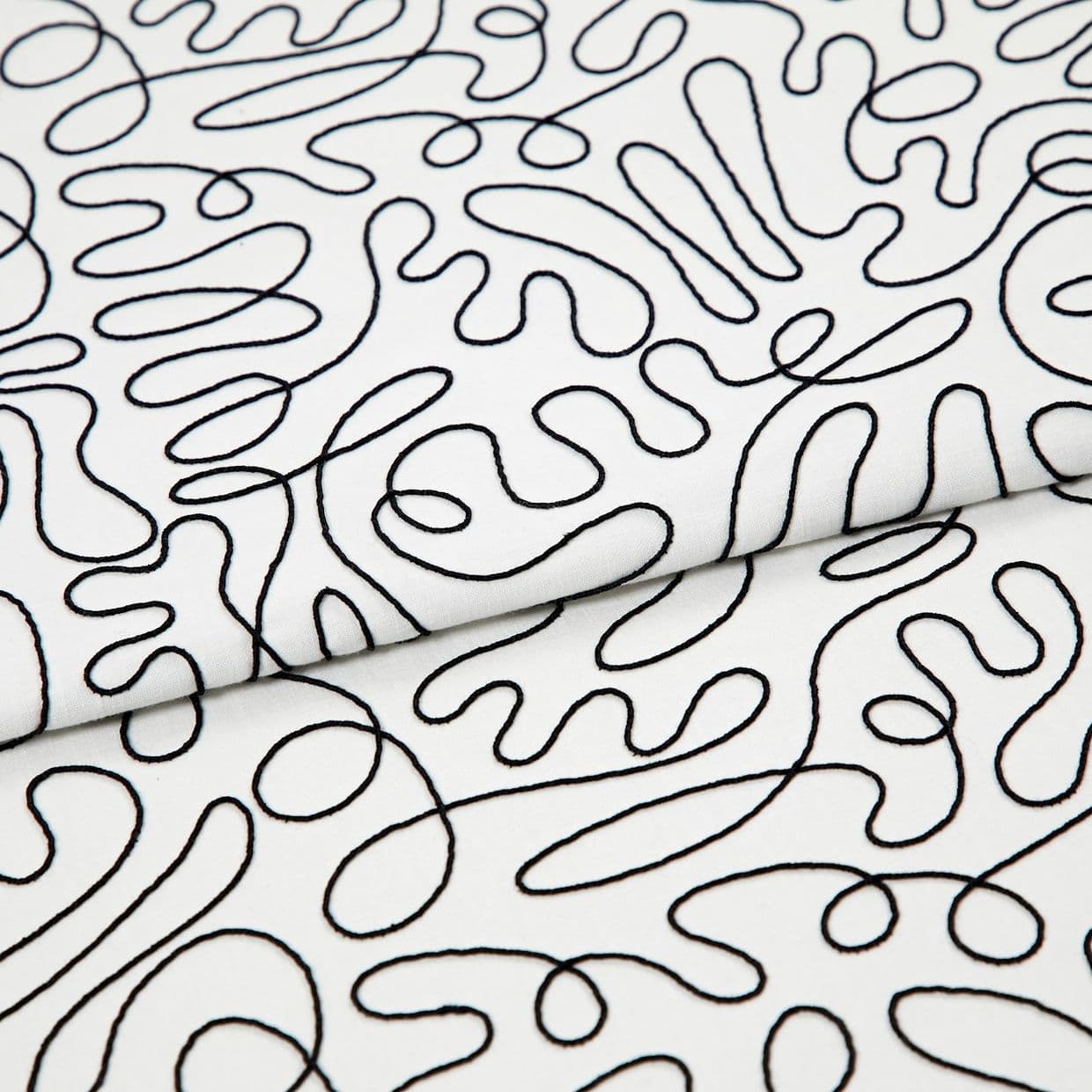 White cotton fabric with continuous black embroidered squiggle pattern, folded once and overlapping; resting flat, showing stitched loops and curves under even studio lighting as a close-up textile sample.