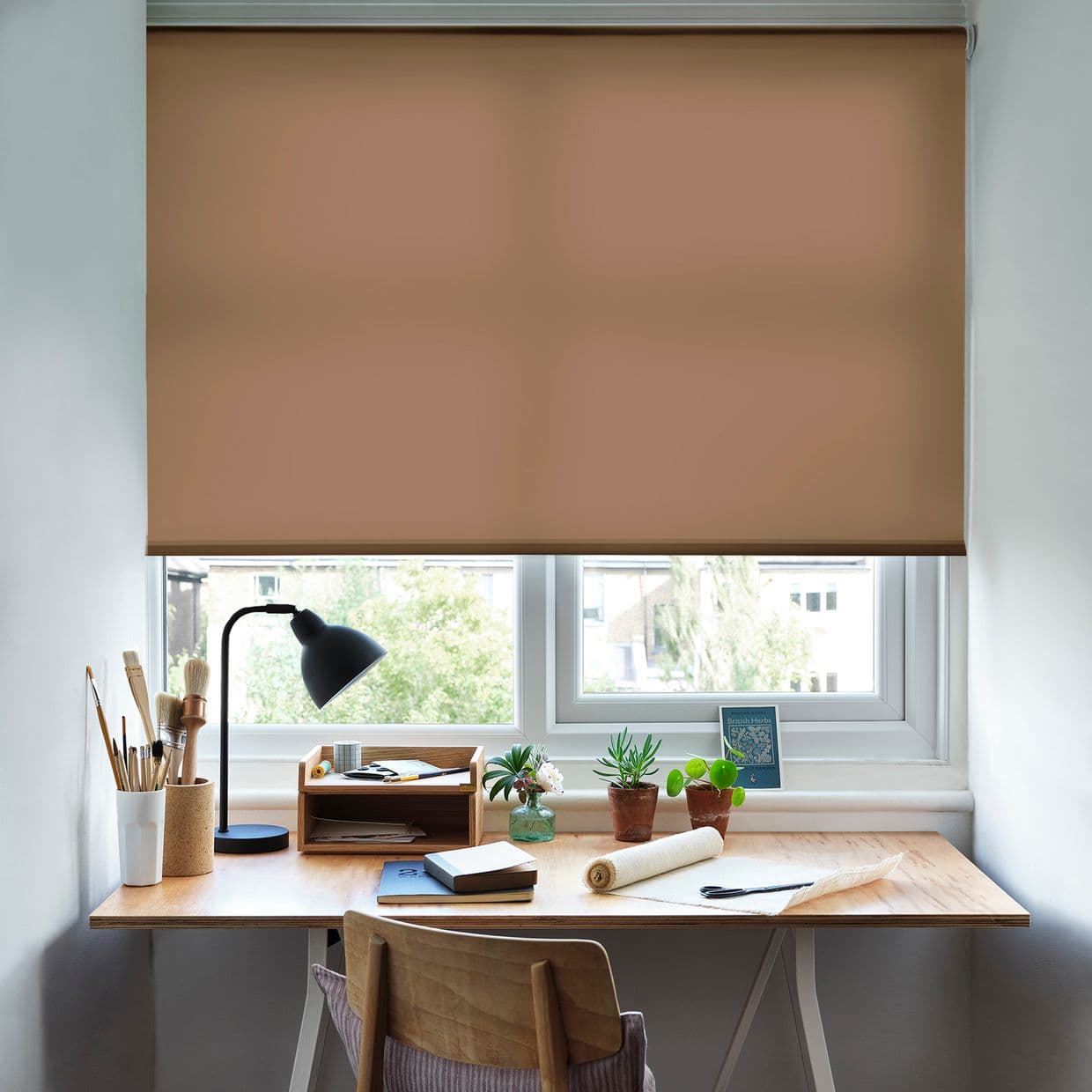 Brown fabric roller blind lowered across a double window, filtering light; wooden desk beneath holds a lamp, brushes, notebooks and small potted plants in a bright home workspace.