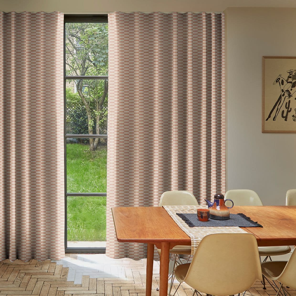 Blush-and-cream checkered full-length curtains, mostly drawn with a narrow central gap; allowing light to frame a mullioned window and green garden, in a sunlit dining room with wooden table.