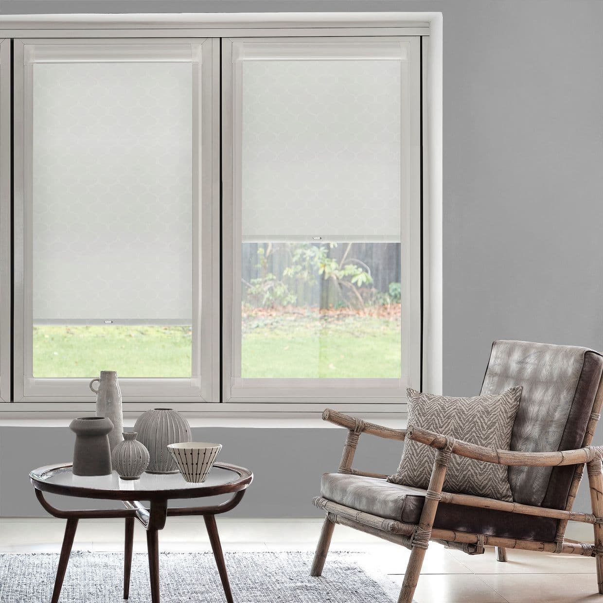 Two white patterned roller blinds, lowered across a double window with an opening on the right, filtering soft daylight; living room with gray walls, rattan armchair and coffee table.
