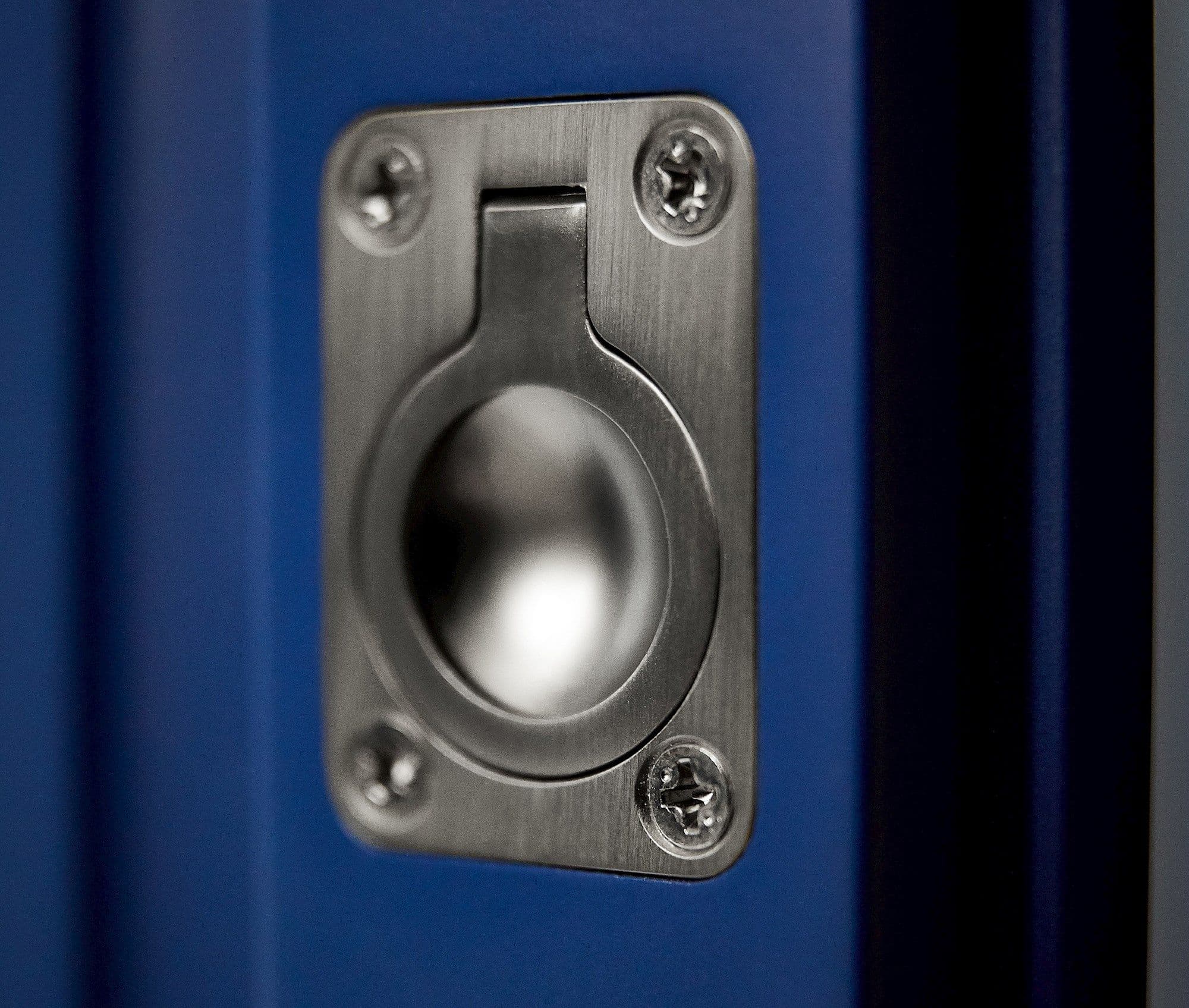 Recessed circular stainless-steel flush pull with rectangular mounting plate and four screws, providing a recessed finger grip; mounted on a blue door, shown close-up with soft, shallow-focused lighting.