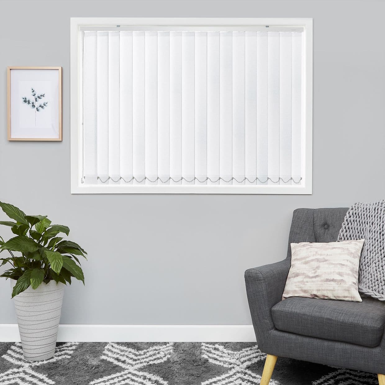 White vertical blinds fully closed over a window, gently filtering daylight; set in a grey living room with a grey armchair, cushion, knit throw, potted plant and framed print.