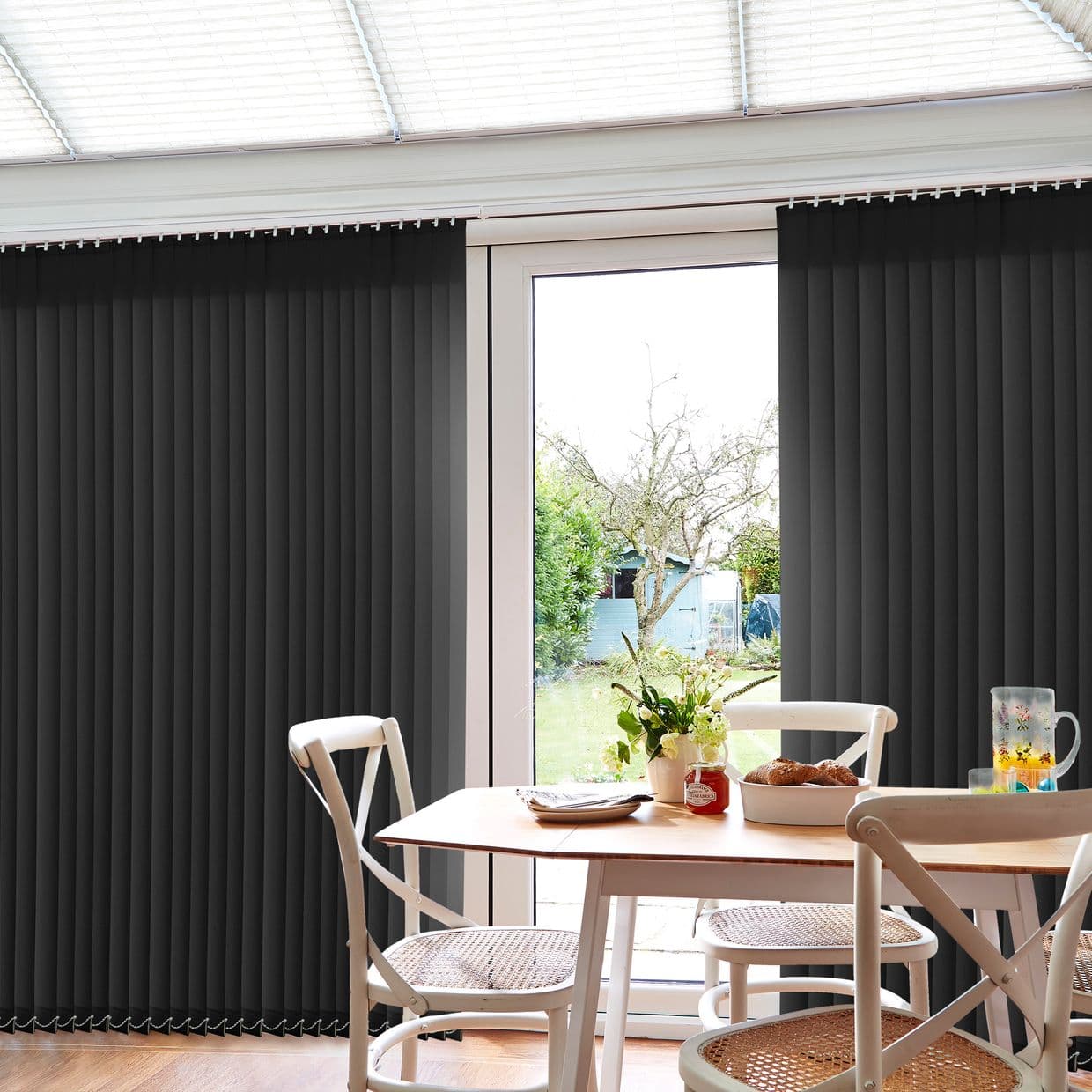Dark vertical blinds cover French doors, parted centrally to expose glass; slats mostly closed, filtering light. Dining table with chairs and vase sits in bright room overlooking a garden.