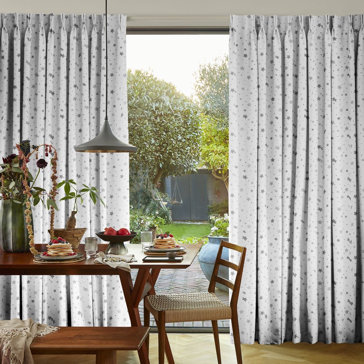 White pleated floor-length curtains with gray star pattern, parted at center to reveal a sunlit garden. They frame a dining table set for breakfast under a pendant lamp.
