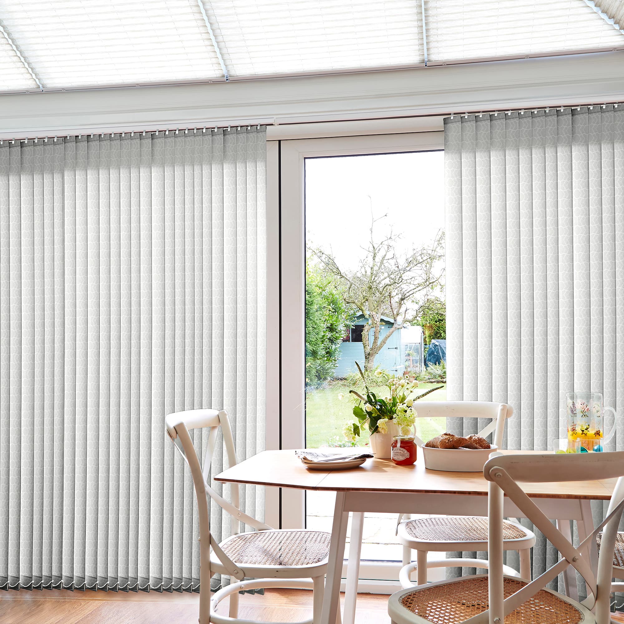 Pale patterned vertical blinds covering a patio door, split open at center to reveal outside; drawn, filtering daylight; small dining table and chairs, vase, garden with tree and shed beyond.