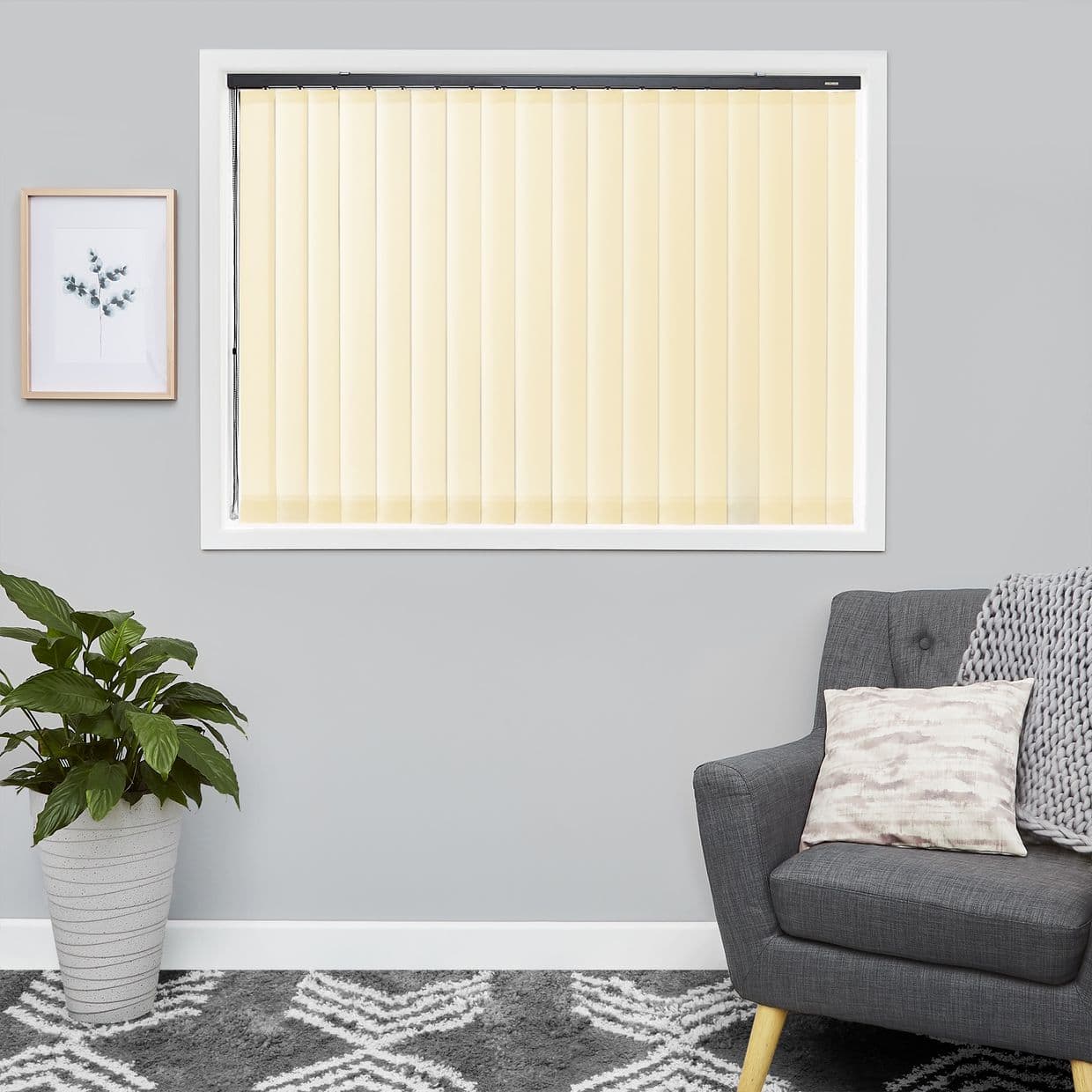 Cream vertical blinds across a central windowâ€”narrow slats on a black headrailâ€”closed, softly filtering daylight, in a grey-walled living room with a potted plant, framed print, patterned rug, and grey armchair.
