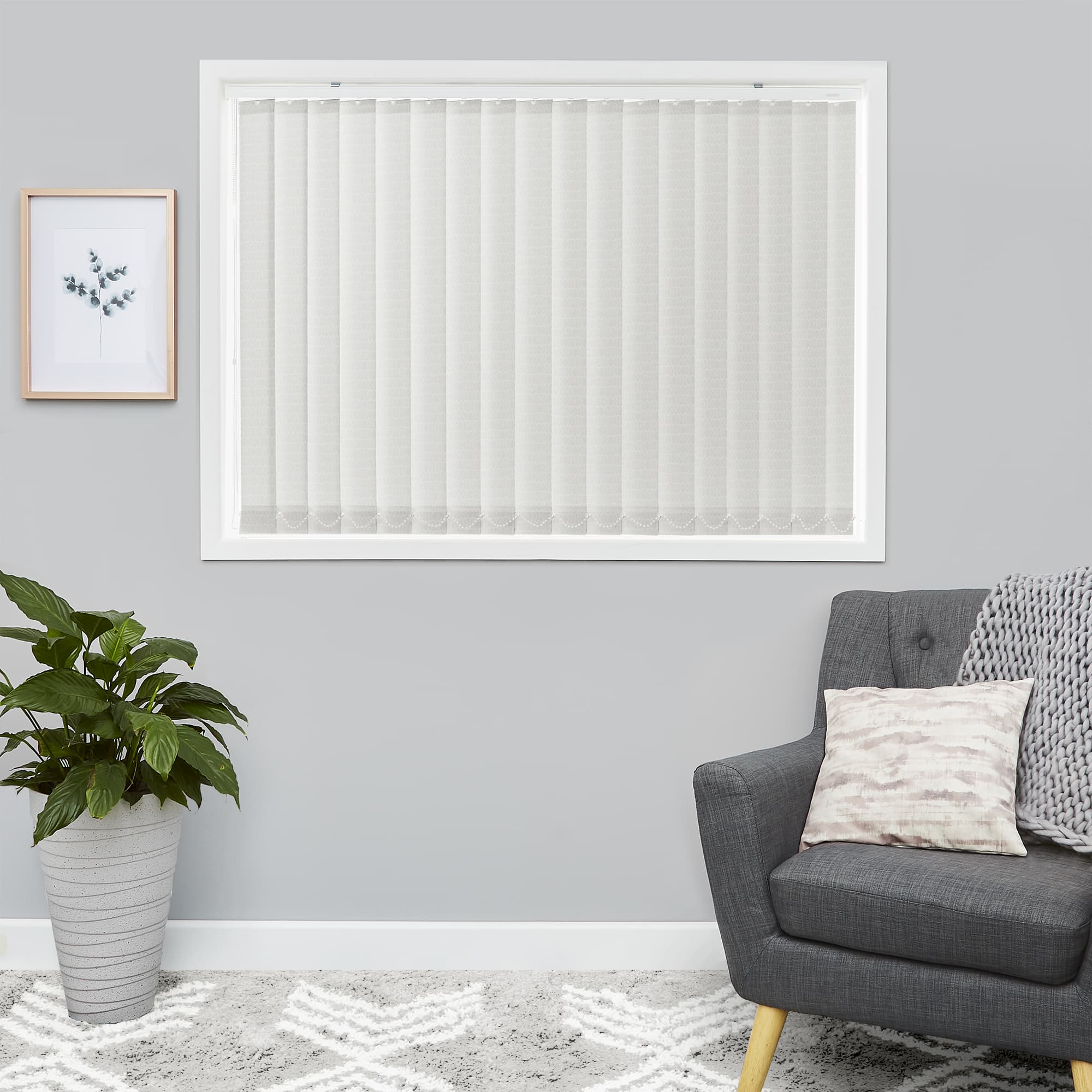 Vertical fabric blinds, closed across a white-framed window, filtering soft daylight into a gray living room with a gray armchair, throw, framed plant print, and potted floor plant.