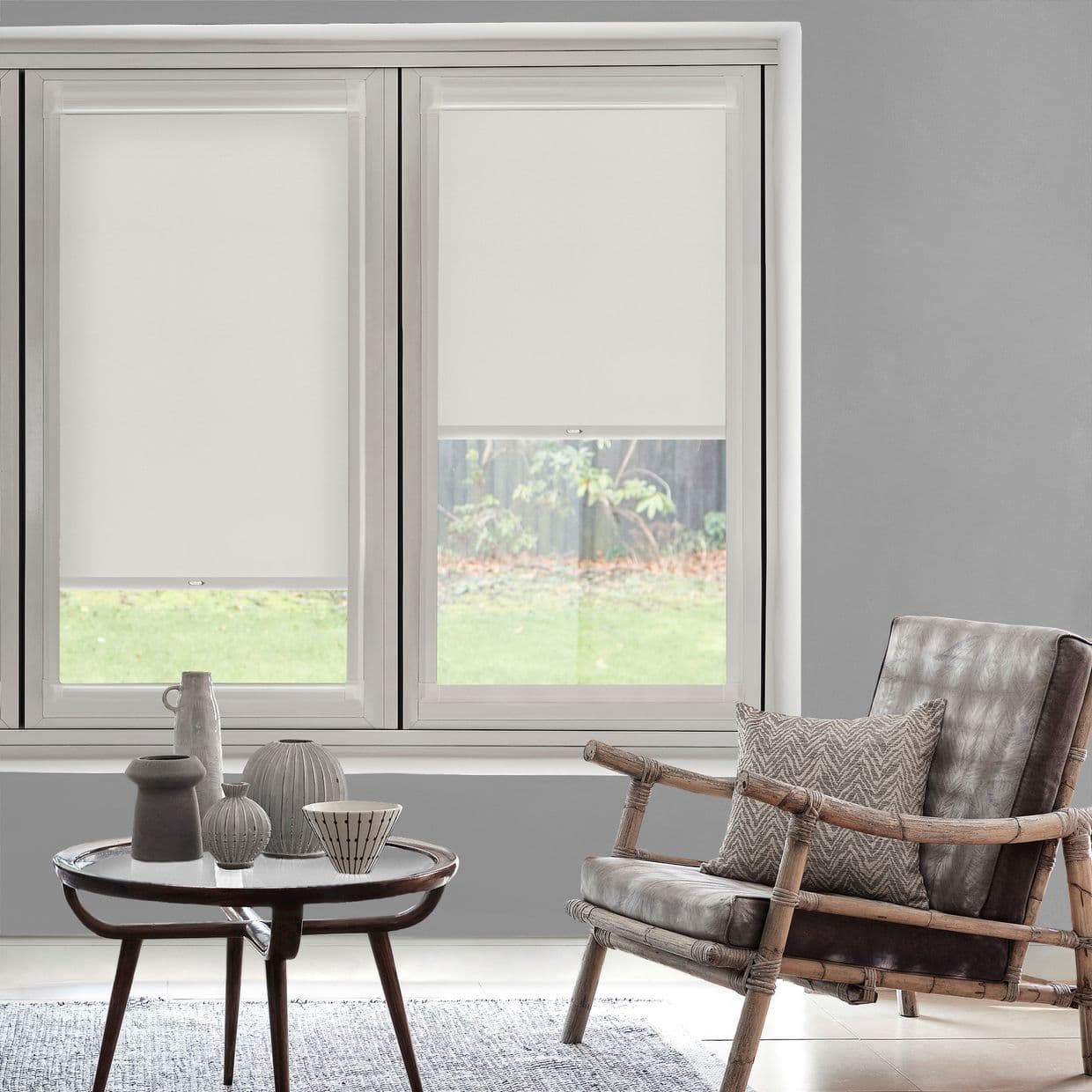 White fabric roller blinds, two-panel, partially lowered covering twin windows with cassette heads, filtering daylight; living area with gray walls, bamboo-frame leather chair, round wooden coffee table, garden view.
