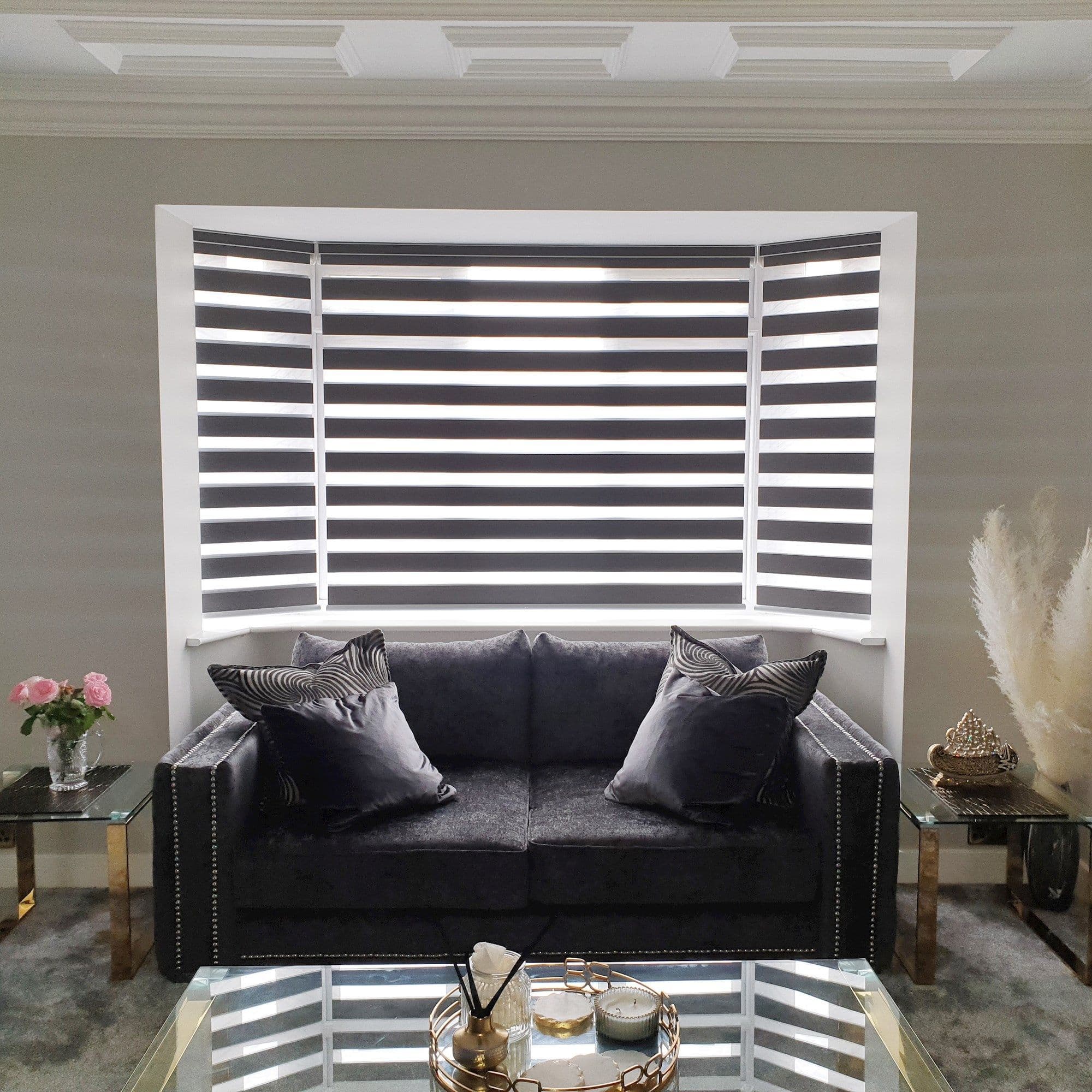 Zebra roller blinds covering a bay window, alternating opaque and sheer horizontal bands, filtering daylight; in a living room with a dark velvet sofa, glass side tables and decorative accents.