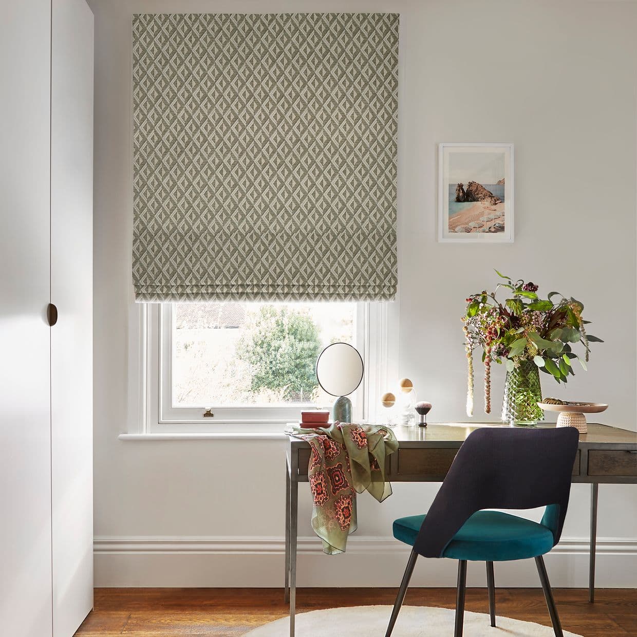 Geometric-patterned Roman blind lowered over a single sash window, gently filtering daylight; small desk beneath with teal chair, vase of flowers, mirror and framed seaside print in a neutral home office.