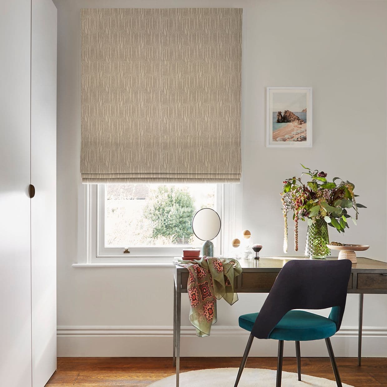 Beige patterned Roman blind, lowered over the window, softly filtering daylight; slim desk below holds round mirror, vase of flowers, scattered accessories, teal-upholstered chair in a neutral home office.