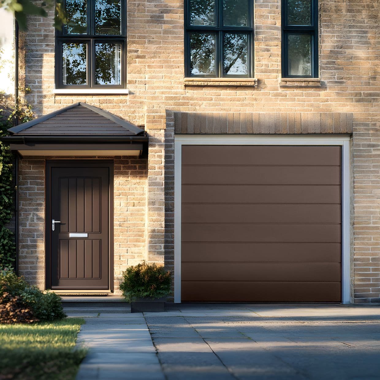 Brown horizontal-panel garage door, closed and flush in a white frame; nearby covered entry door and upper black-framed windows reflect trees, sunlit brick faÃ§ade and a tidy suburban driveway.